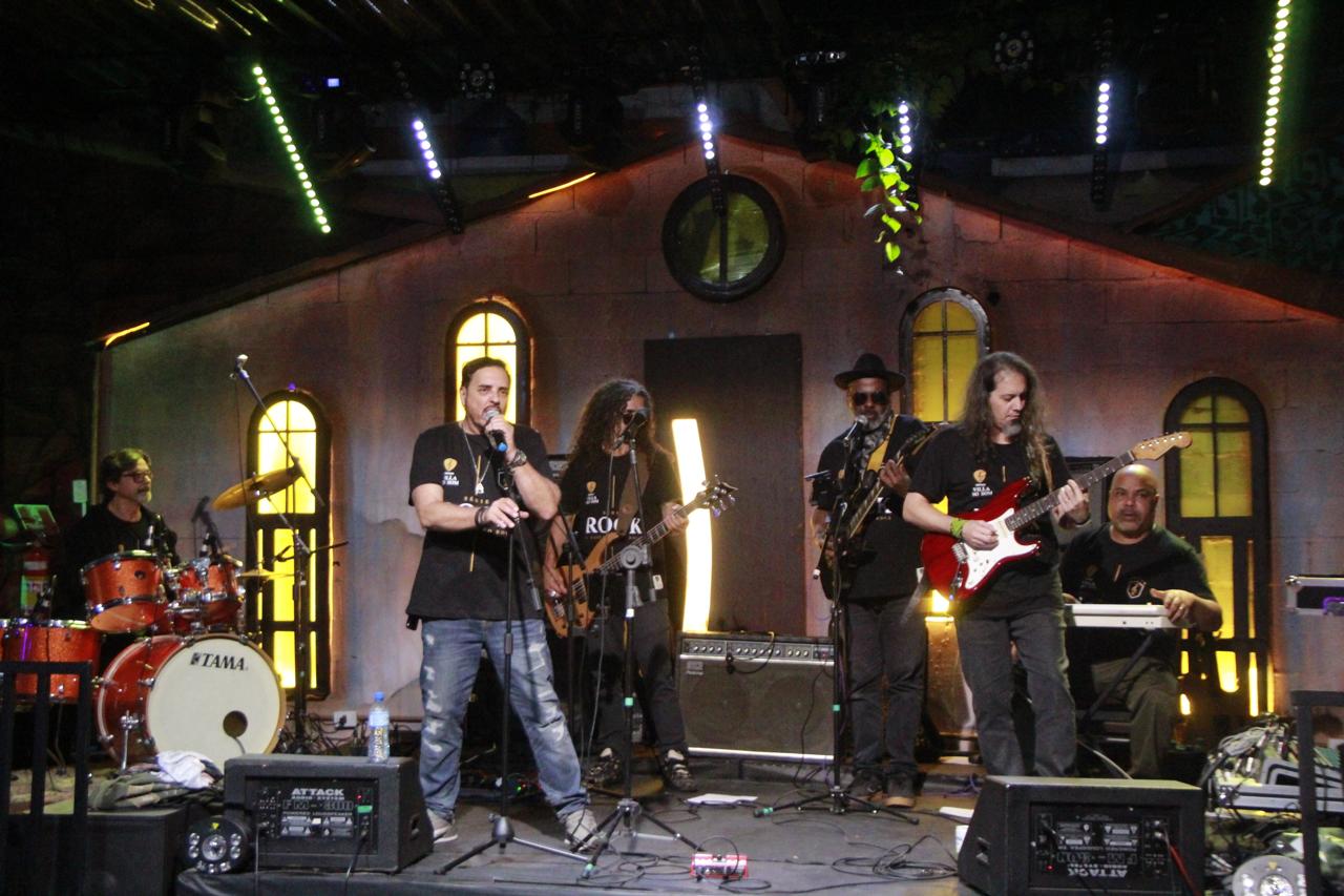 Banda de rock completa no palco cenográfico da Villa do Som durante show ao vivo com iluminação verde neon e rig profissional